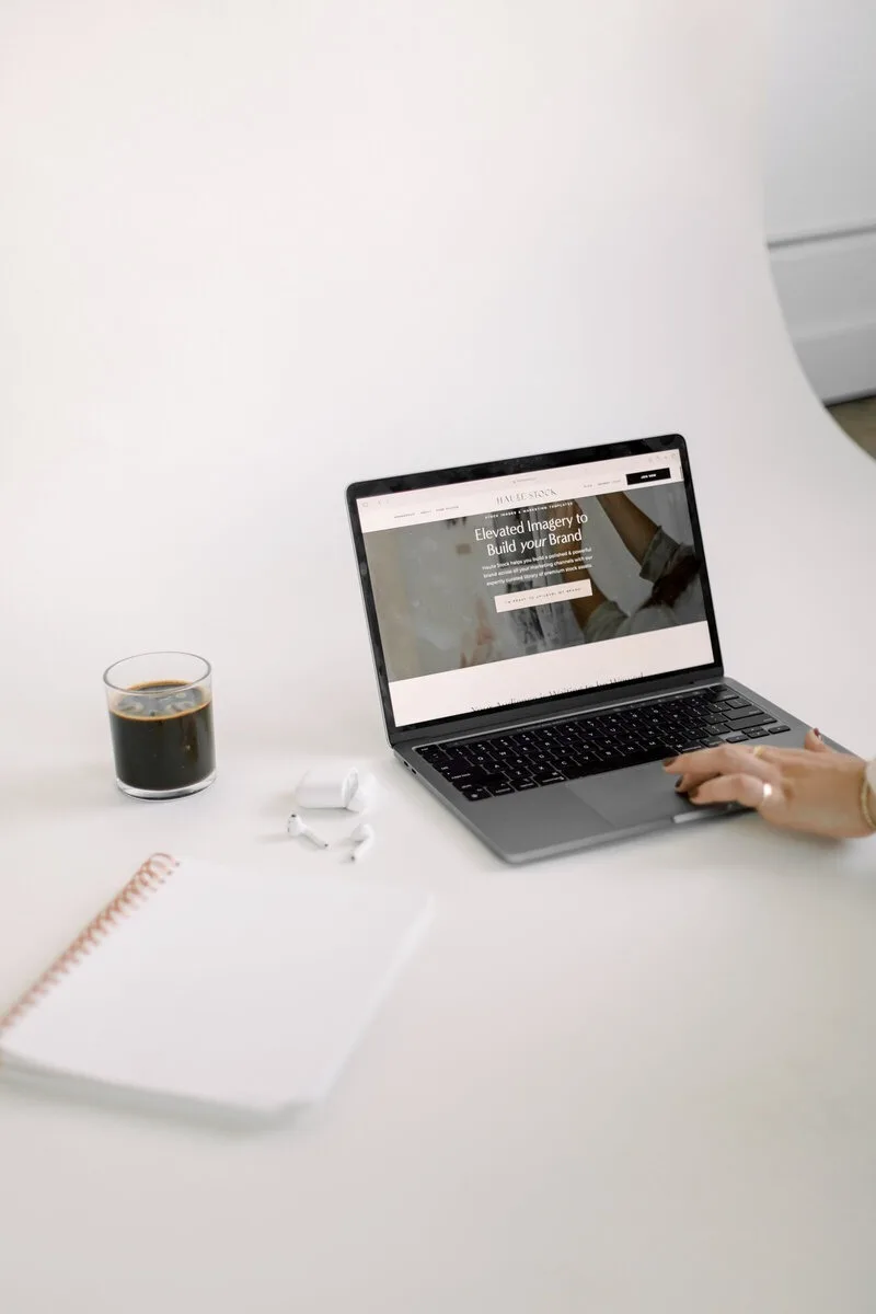 Laptop on a white desk beside coffee, a notebook, and earbuds.