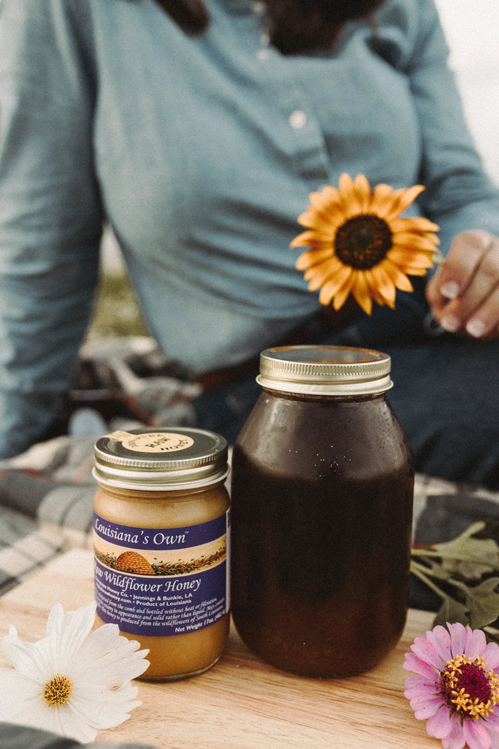 Two jars of honey sit on a wooden surface with flowers and a seated person in the background.