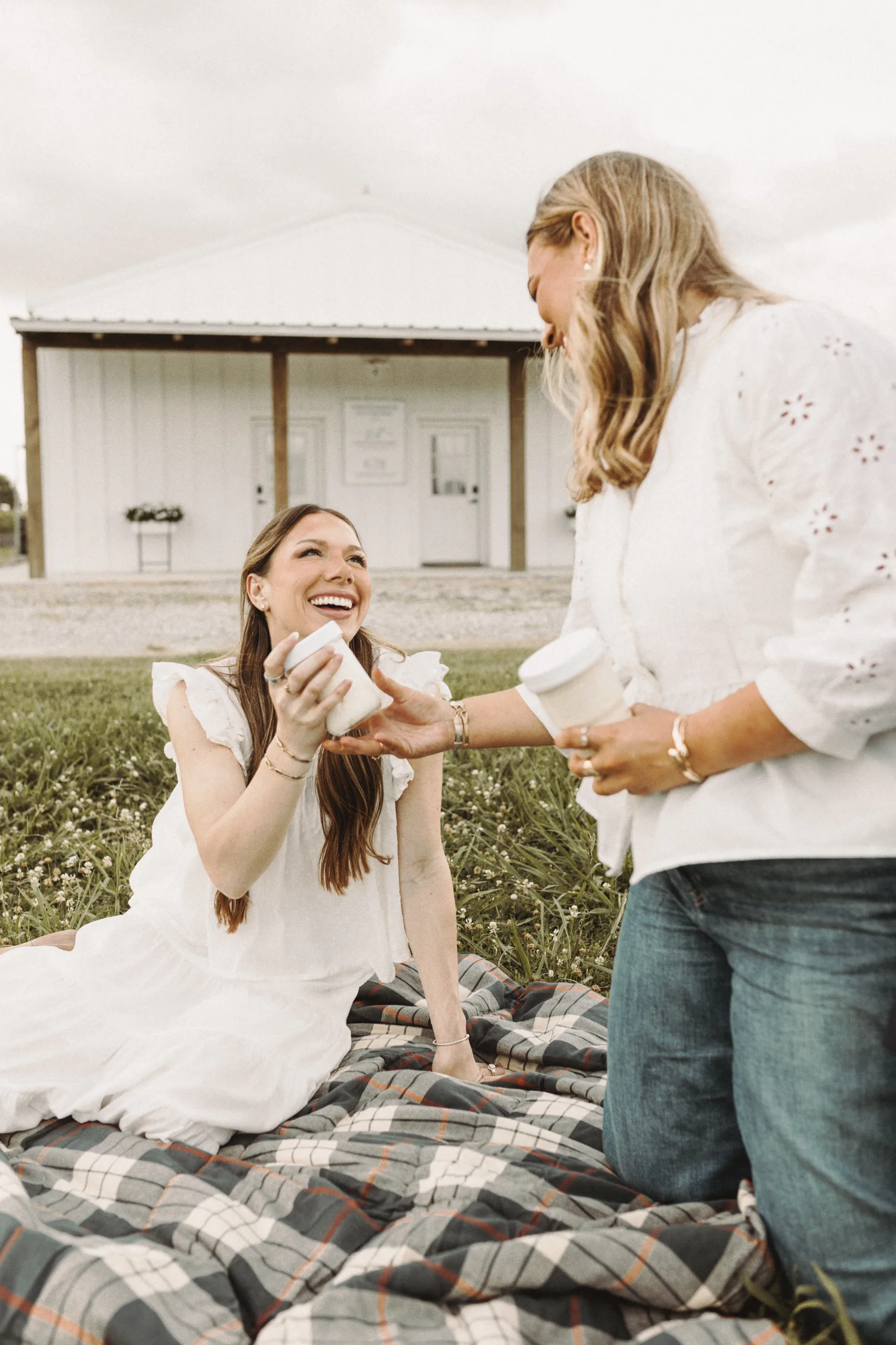 Two women share a drink on a blanket beside a grassy field.